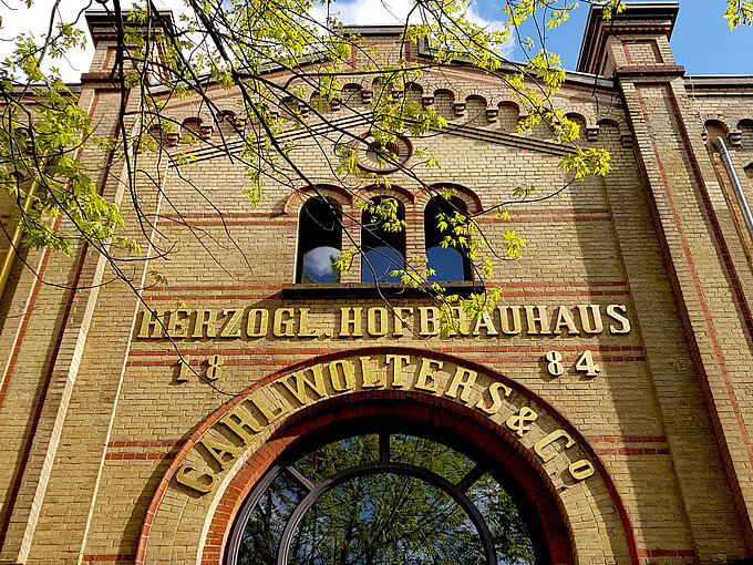Backstein-Fassade des Herzog Hofbrauhauses mit goldener Schrift unter blauen Himmel und frischen grünen Blättern.