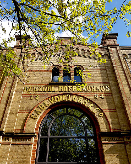 Backstein-Fassade des Herzog Hofbrauhauses mit goldener Schrift unter blauen Himmel und frischen grünen Blättern.