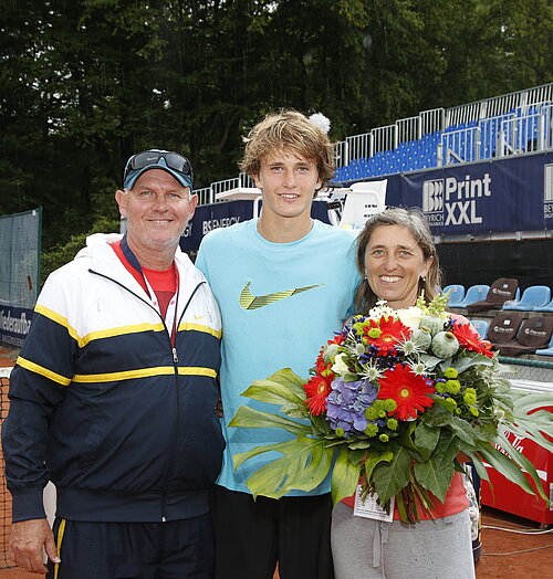 Auf dem Bild sieht man einen jungen blonden Mann mit einem hellblauen Sport-Shirt. Links von ihm steht ein älterer Mann mit Cap und Trainingsjacke. Rechts von ihm steht eine Frau, die einen großen Blumenstrauß in der Hand hält. Im Hintergrund sieht man einen Teil des Tennisplatzes und der Tribünen.