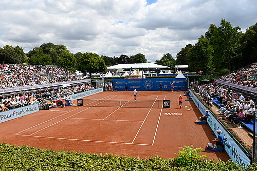 Auf dem Bild sieht man einen großen Tennisplatz mit rotem Sand. Links und rechts vom Platz sind Tribünen mit vielen Menschen.