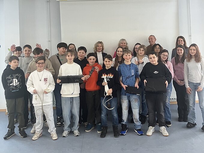 Eine Gruppe von Schülern und Lehrern posiert gemeinsam vor einer weißen Leinwand. Die Schüler halten zum Teil elektronische Gegenstände, wie Tastaturen, Fernbedienungen oder Mehrfachsteckdosen in den Händen.
