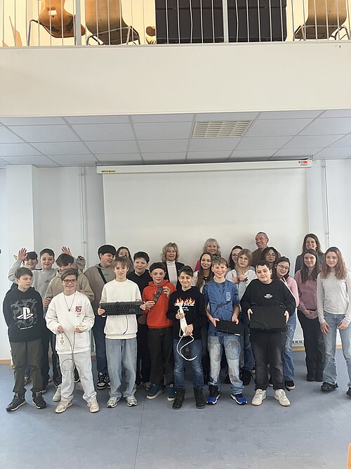 Eine Gruppe von Schülern und Lehrern posiert gemeinsam vor einer weißen Leinwand. Die Schüler halten zum Teil elektronische Gegenstände, wie Tastaturen, Fernbedienungen oder Mehrfachsteckdosen in den Händen.