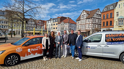 Gruppe von sieben Personen posiert vor zwei Autos in der malerischen Altstadt von Peine mit Fachwerkhäusern.