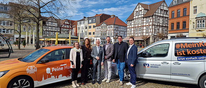 Gruppe von sieben Personen posiert vor zwei Autos in der malerischen Altstadt von Peine mit Fachwerkhäusern.