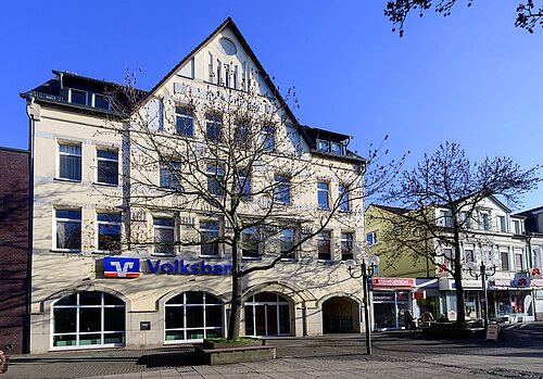 Fassade eines historischen Gebäudes mit Volksbank-Logo, umgeben von Bäumen, bei klarem Himmel.