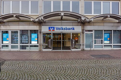 Außenansicht einer Volksbank-Filiale mit einem modernen Eingang. Über der Tür befindet sich das Volksbank-Logo. Die Fenster links und rechts sind mit Plakaten dekoriert, die auf Aktionen hinweisen. Der Gehweg davor ist mit Pflastersteinen ausgelegt.