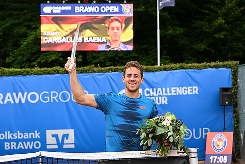 Auf dem Bild sieht man einen braunhaarigen Mann mit einem blauen Sportshirt vor einer blauen Werbewand. In seiner rechten Hand hält er einen transparenten Pokal hoch und in der anderen Hand einen Blumenstrauß.