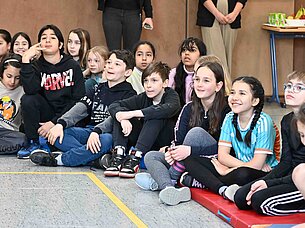 Kinder sitzen auf dem Boden einer Turnhalle, lächeln und beobachten etwas mit Interesse. Im Hintergrund steht ein Tisch.