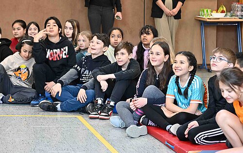Kinder sitzen auf dem Boden einer Turnhalle, lächeln und beobachten etwas mit Interesse. Im Hintergrund steht ein Tisch.