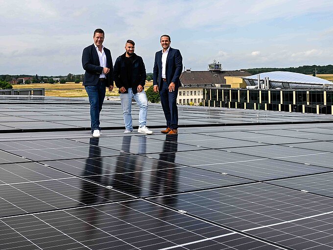 Drei Männer stehen auf einem Solarzellen-Dach mit Landschaft im Hintergrund, Himmel leicht bewölkt.