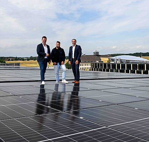 Drei Männer stehen auf einem Solarzellen-Dach mit Landschaft im Hintergrund, Himmel leicht bewölkt.