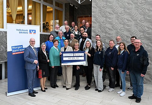 Gruppe von Menschen überreicht Scheck von 46.024,72 Euro vor einem Gebäude, neben einem Volksbank-Banner.