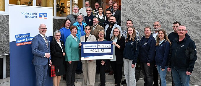 Gruppe von Menschen überreicht Scheck von 46.024,72 Euro vor einem Gebäude, neben einem Volksbank-Banner.