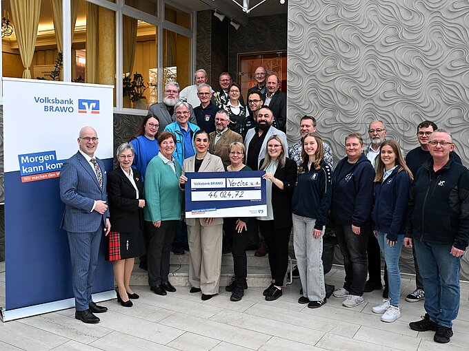 Gruppe von Menschen überreicht Scheck von 46.024,72 Euro vor einem Gebäude, neben einem Volksbank-Banner.