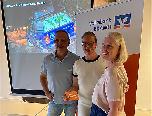 Drei lächelnde Personen (Joachim Franz (Referent), Nicole Mölling und Stephanie Borrmann) stehen nebeneinander vor einem Präsentationsbildschirm und einem Roll-Up mit dem Logo der „Volksbank BRAWO“. Im Hintergrund zeigt der Bildschirm ein Bild eines bunt beklebten Geländewagens mit der Aufschrift „Angst – den Weg nicht zu finden.“ Die mittlere Person hält eine orangefarbene Karte mit dem Text „Willst Du mit mir gehen?“ in der Hand.