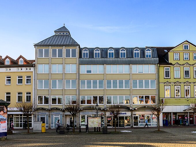 Städtische Häuserfassade mit Geschäften, kahlen Bäumen und einer Informationstafel auf einem sonnenbeschienenen Platz.