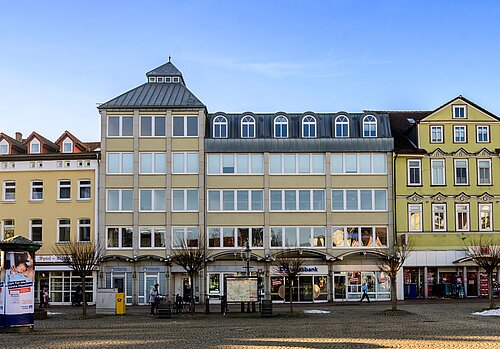 Städtische Häuserfassade mit Geschäften, kahlen Bäumen und einer Informationstafel auf einem sonnenbeschienenen Platz.