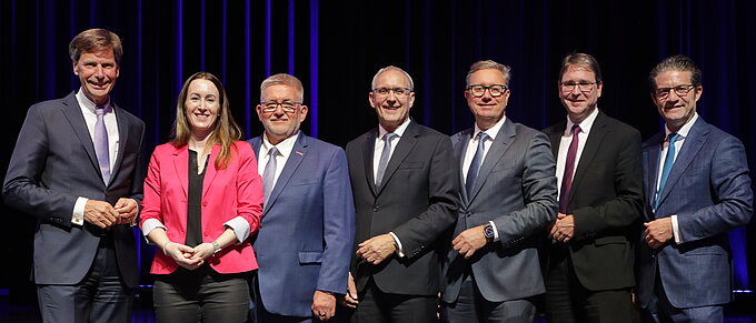 Gruppe von sieben Personen in Anzügen steht auf einer Bühne und lächelt in die Kamera.