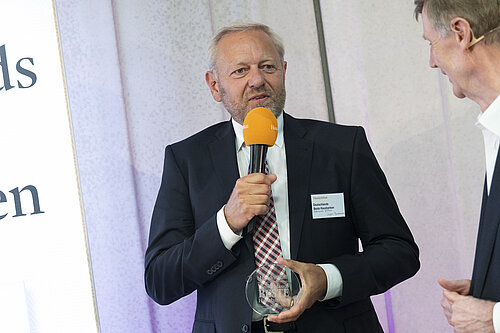 Ein Mann in einem Anzug hält ein Mikrofon und eine Auszeichnung. Er steht vor einem weißen Hintergrund mit Text und spricht scheinbar auf einer Veranstaltung. Neben ihm steht eine weitere Person mit einem weißen Hemd. Die Szene wirkt förmlich und professionell.