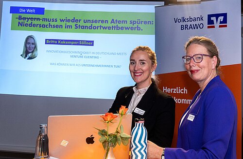 Auf dem Bild sieht man zwei Frauen vor einer Präsentation. Die linke Frau trägt einen schwarzen Blazer und die rechte Frau einen blauen Blazer. Beide haben die Haare zu einem Dutt gebunden und lächeln in die Kamera.