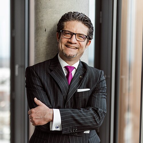 Lächelnder Mann im Anzug mit Brille vor einem Fenster stehend, mit verschränkten Armen.