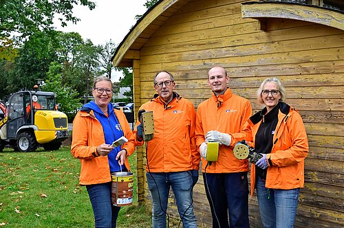 Vier Personen in orangefarbenen Jacken stehen vor einem Holzschuppen. Sie halten Werkzeuge, wie Pinsel und Schwingschleifer, in den Händen. Im Hintergrund ist ein gelber Radlader und grüne Bäume zu sehen. Alle lächeln in die Kamera.