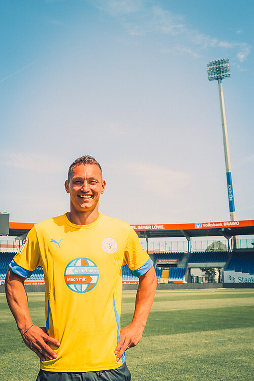 Ein junger Mann in einem gelben Fußballtrikot mit blauen Akzenten steht auf einem Fußballfeld und lächelt in die Kamera. Im Hintergrund sind leere Tribünen und ein hoher Flutlichtmast zu sehen. Auf dem Trikot sind das Logo von Puma, das Vereinswappen von Eintracht Braunschweig und das Sponsorenlogo "walk4help – Mach mit!" zu erkennen.