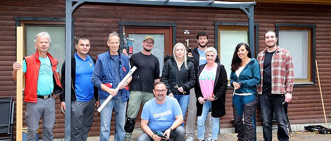 Eine Gruppe von zehn Personen steht vor einem hölzernen Gebäude. Sie sind unterschiedlich gekleidet, einige halten Werkzeuge wie Holzlatten, eine Wasserwaage und einen Bohrer in den Händen. Die Stimmung wirkt freundlich und kooperativ. Die Fassade des Gebäudes ist mit Fenstern versehen, und das Dach ist mit Moos bewachsen.