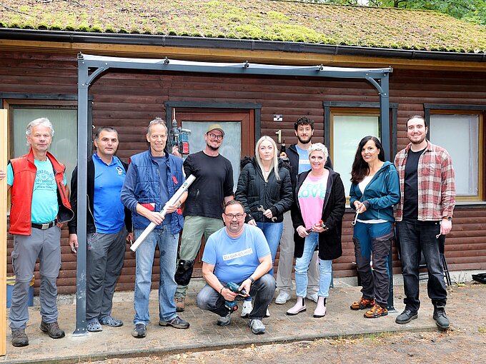 Eine Gruppe von zehn Personen steht vor einem hölzernen Gebäude. Sie sind unterschiedlich gekleidet, einige halten Werkzeuge wie Holzlatten, eine Wasserwaage und einen Bohrer in den Händen. Die Stimmung wirkt freundlich und kooperativ. Die Fassade des Gebäudes ist mit Fenstern versehen, und das Dach ist mit Moos bewachsen.