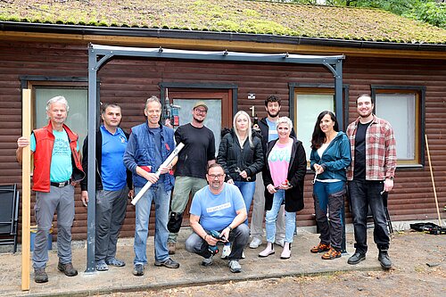 Eine Gruppe von zehn Personen steht vor einem hölzernen Gebäude. Sie sind unterschiedlich gekleidet, einige halten Werkzeuge wie Holzlatten, eine Wasserwaage und einen Bohrer in den Händen. Die Stimmung wirkt freundlich und kooperativ. Die Fassade des Gebäudes ist mit Fenstern versehen, und das Dach ist mit Moos bewachsen.