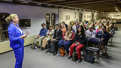 Eine Frau in einem blauen Anzug hält einen Vortrag vor einem weiblichen, sitzenden Publikum in einem Raum mit Fotografien an den Wänden.