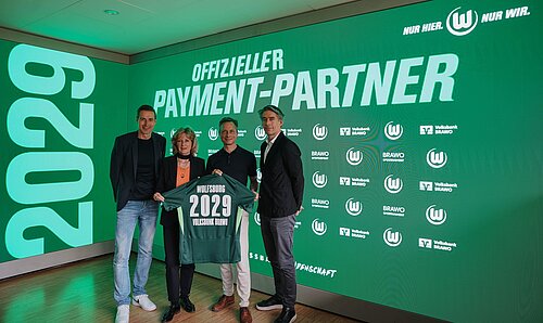 Vier Personen stehen vor einer LED-Leinwand und halten ein Trikot in der Hand, das u. a. mit der Zahl 2029 beflockt ist..