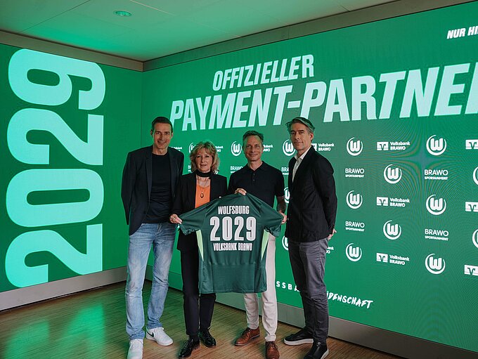 Vier Personen stehen vor einer LED-Leinwand und halten ein Trikot in der Hand, das u. a. mit der Zahl 2029 beflockt ist..