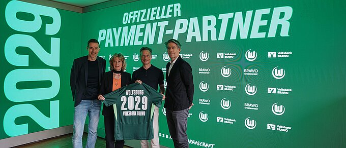 Vier Personen stehen vor einer LED-Leinwand und halten ein Trikot in der Hand, das u. a. mit der Zahl 2029 beflockt ist..