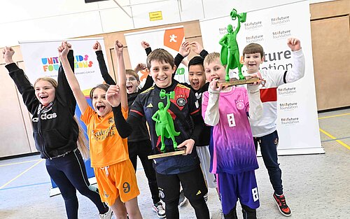 Kinder jubeln gemeinsam in Sporttrikots und halten grüne Trophäen vor fit4future Foundation Bannern.