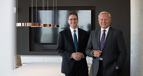 Zwei Männer in Anzügen stehen in einem modernen Büro mit einer großen Bildschirmpräsentation im Hintergrund.
