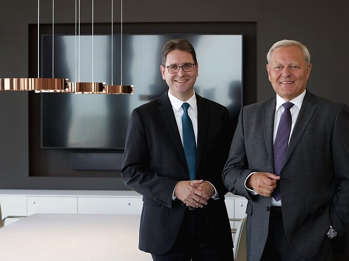Zwei Männer in Anzügen stehen in einem modernen Büro mit einer großen Bildschirmpräsentation im Hintergrund.