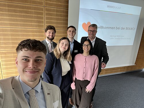 Gruppe von sechs Personen posiert lächelnd vor einer Präsentationstafel mit der Aufschrift "Willkommen bei der BRAWO!"