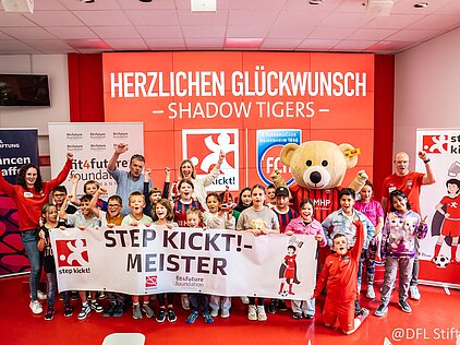Gruppe von Kindern und Erwachsenen feiert den „Step Kickt!“-Meistertitel mit Maskottchen und Bannern im Hintergrund.