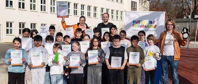 Gruppe von Kindern und Erwachsenen posiert mit Zertifikaten und Plüschtieren vor einem Schulgebäude.