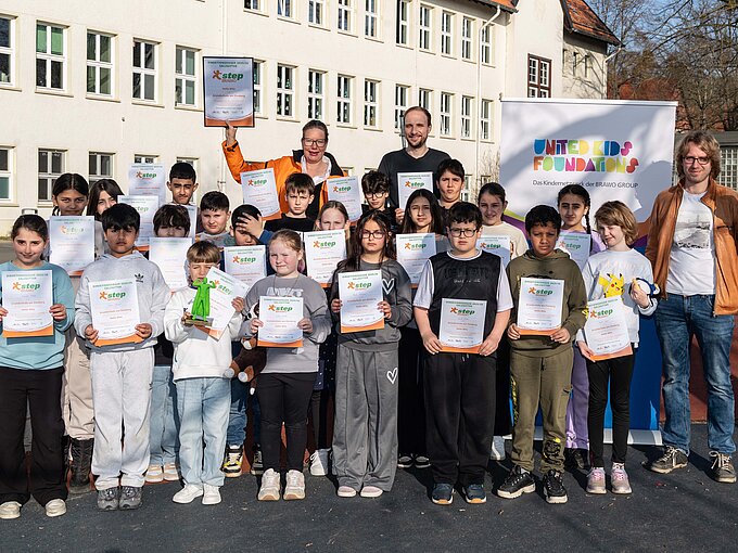Gruppe von Kindern und Erwachsenen posiert mit Zertifikaten und Plüschtieren vor einem Schulgebäude.