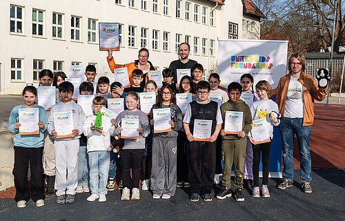 Gruppe von Kindern und Erwachsenen posiert mit Zertifikaten und Plüschtieren vor einem Schulgebäude.