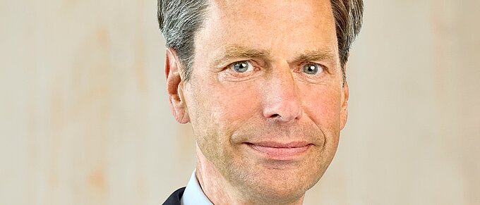 Mann in Anzug und Krawatte vor neutralem Hintergrund, schaut in die Kamera.