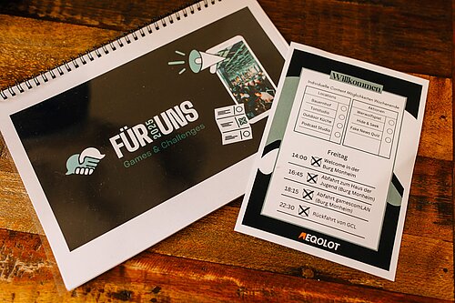 Notizbuch und Plan mit Events und Aktivitäten, inklusive Games & Challenges, auf einem Holztisch.