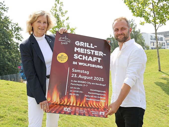 Zwei Personen stehen und halten ein Plakat fest zur Ankündigung der Veranstaltung.