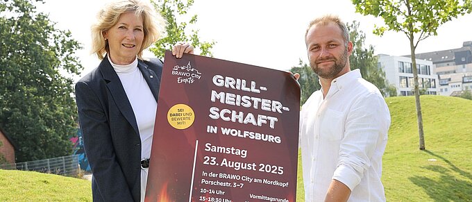 Zwei Personen stehen und halten ein Plakat fest zur Ankündigung der Veranstaltung. 
