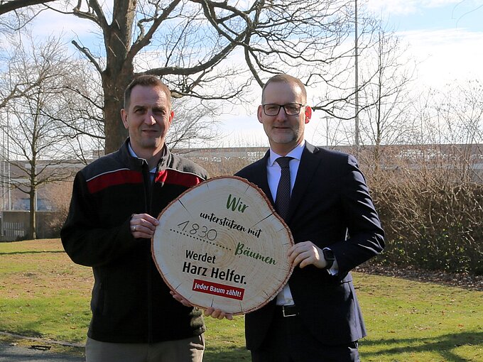Zwei Männer halten ein großes rundes Holzschild mit der Aufschrift "Wir unterstützen mit 1.830 Bäumen. Werdet Harz Helfer. Jeder Baum zählt!"