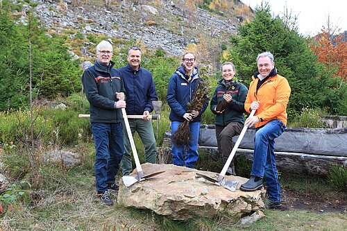 Fünf Personen stehen mit Spaten und Setzlingen in einem bewaldeten Gebiet.