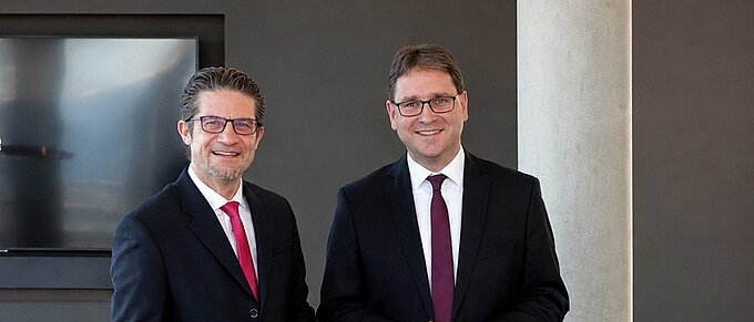 Zwei Männer in Anzügen stehen in einem modernen Büro, lächelnd, vor einem Bildschirm und einer Betonwand.
