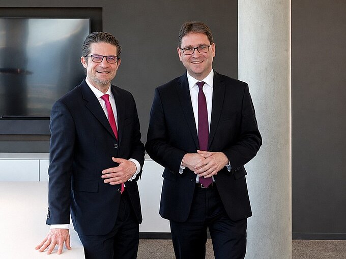 Zwei Männer in Anzügen stehen in einem modernen Büro, lächelnd, vor einem Bildschirm und einer Betonwand.
